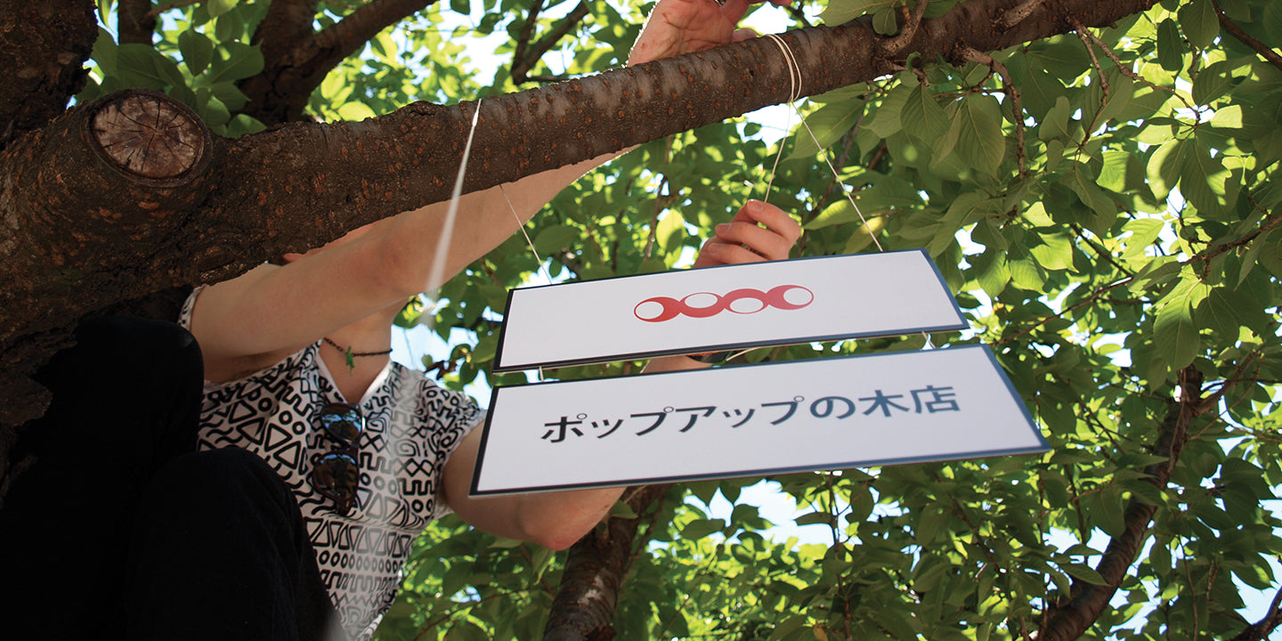 birds eye view of Duncan tying a paper sign onto a tree branch. The sign reads Pop up Tree Shop in Japanese