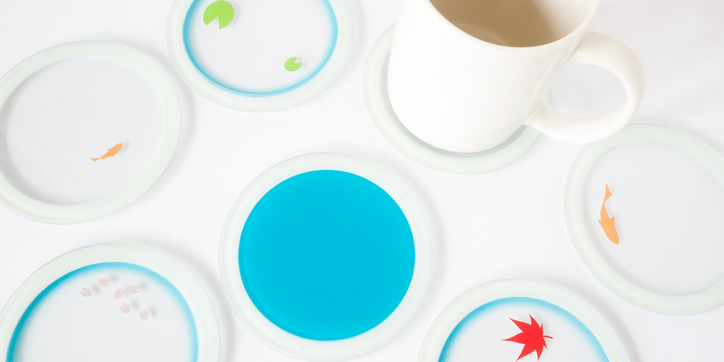 goldfish bowl glass coasters laid out on a surface, showing goldfish and frosted bowl walls, and a ceramic white mug cup