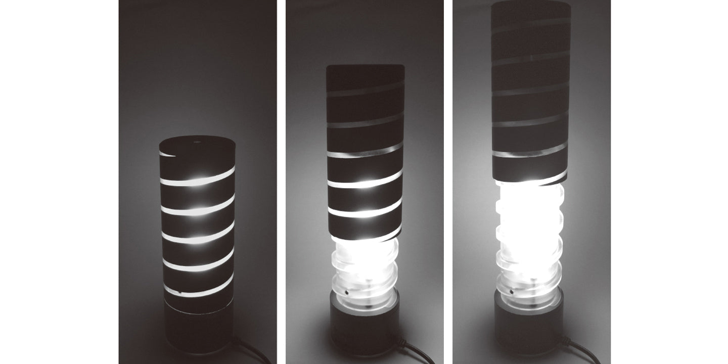 Desk light that has a clear lamp shade in a spiral, screw-like shape, and a secondary cover that is mostly opaque and can screw up and down the lamp shade.
Room light shown in three stages of operation; First, fully twisted in closed position, only releasing minimal light through the small gaps in the cover. Then half twisted off, with medium light released, and finally in maximum open position, releasing the most amount of light